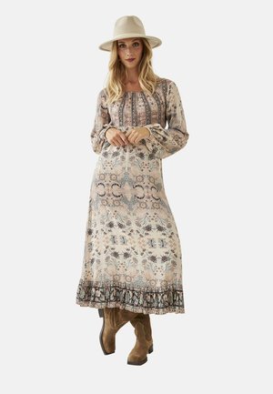 Mujer con un vestido largo de estampado floral y mangas abullonadas, sombrero de ala ancha color marrón claro y botas de ante marrones, posando frente a un fondo liso.