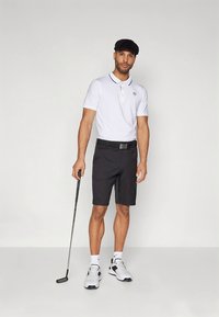 Pánsky golfový outfit so bielym polokošele s čiernym golierom, čiernymi šortkami, opaskom a bielymi teniskami so zelenými detailmi. Drží golfovú palicu.