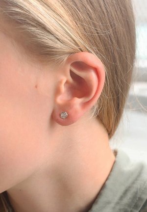 Zilveren bloemvormige oorbel met een heldere edelsteen in het midden, afgebeeld tegen een lichte huidtint en lang blond haar.