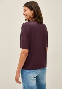 Top morado de manga corta con un corte relajado, que presenta detalles de botones en los hombros, hecho de una tela suave. Combinado con jeans azul claro.