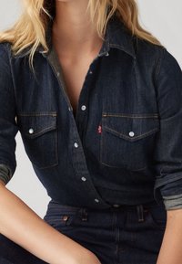 Mörk denimskjorta med knappstängning framtill, två bröstfickor och kontrasterande sömmar. Tyget har en slät yta och en väldefinierad passform.