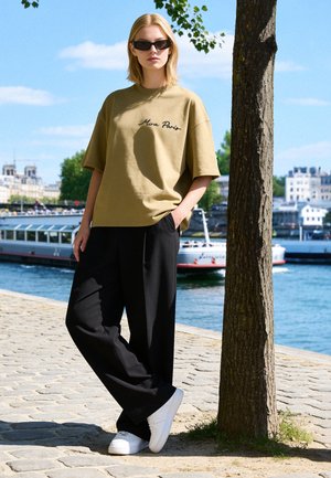Donna con occhiali da sole, camicia beige con testo "Mira Paris", pantaloni neri larghi e scarpe bianche, appoggiata a un albero vicino a un fiume con barche.