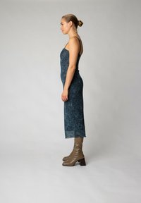 O rochie mulată, de culoare teal, cu un model geometric, bretele subțiri și margine zimțată, asortată cu cizme bej cu toc până la genunchi.