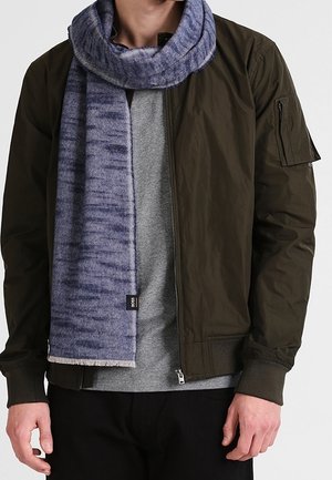 Homme portant une chemise grise, une veste bomber vert olive, un pantalon noir et une écharpe bleue à motifs enroulée autour du cou.