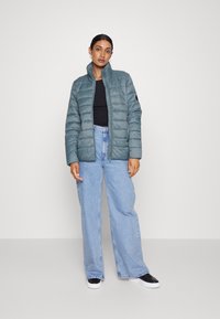 Casaco puffer azul claro com colarinho alto, design acolchoado e fecho de correr. Combinado com jeans de perna larga azul claro e tênis pretos.