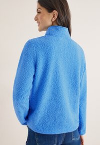 Femme aux cheveux bruns portant une veste en polaire duveteuse bleu vif et un jean bleu foncé, vue de dos et souriante, sur un fond uni.