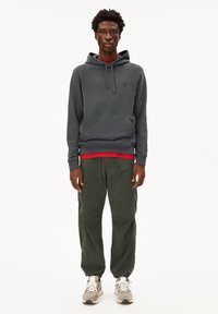 Felpa grigia con cappuccio e tasca frontale, indossata sopra una maglietta rossa. I pantaloni sportivi verde oliva e le scarpe da ginnastica grigie completano l'outfit casual.