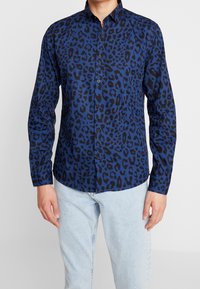 Niebieska koszula w panterkę z kołnierzykiem na guziki, długimi rękawami, czarnymi akcentami i gładką fakturą. W połączeniu z jasnymi dżinsami.