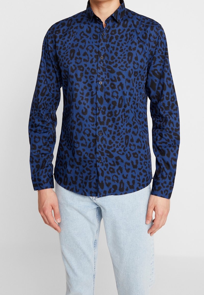 Niebieska koszula w panterkę z kołnierzykiem na guziki, długimi rękawami, czarnymi akcentami i gładką fakturą. W połączeniu z jasnymi dżinsami.