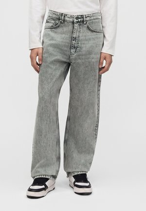 Person, die lockere graue Jeans mit Waschung und schwarz-weiße Sneakers trägt, steht vor einem einfachen hellen Hintergrund.