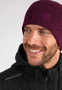 Gorro de punto burdeos con un patrón sutil y logo bordado; lo lleva puesto un hombre con una sudadera negra con cremallera y un acabado texturizado.