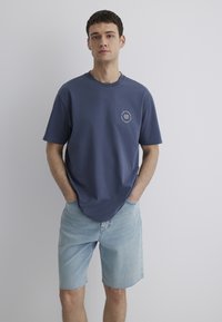 Navy blue cotton t-shirt with short sleeves, featuring a circular graphic design on the chest, paired with light blue denim shorts.
