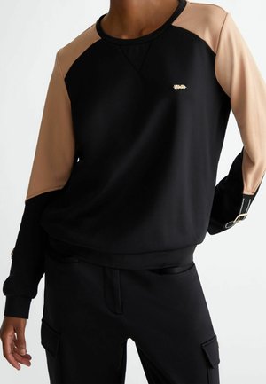 Person wearing black sweatshirt with beige raglan sleeves and gold buckle details, paired with black cargo pants.