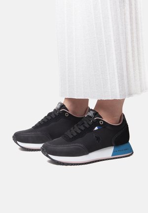 Sorte U.S. Polo Assn. sneakers med blå hælaccents båret med et hvidt plisseret skørt, vist fra siden på en ensfarvet baggrund.