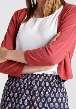 Femme les bras croisés portant un top blanc, un cardigan court corail et une jupe bleu marine à motif floral sur un fond clair.