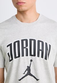 Grå bomulds T-shirt med fed sort "JORDAN" tekst og en stiliseret sort silhuet af en springende figur med en basketball.