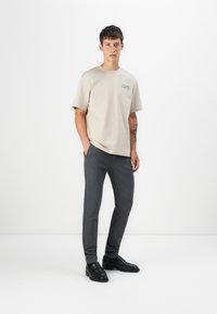 Beige bomull T-skjorte med korte ermer og en liten blå logo, kombinert med mørkegrå slim-fit bukser og svarte sko. Glatt tekstur.