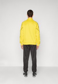 Gul sweatshirt med høj krave, sort piping langs sømmene, parret med mørke sweatpants og sorte atletiske sko.