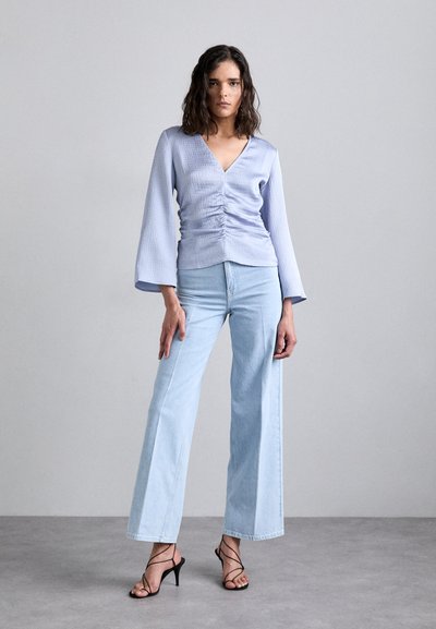 Lyseblå, teksturert topp med V-hals og rynkedesign; parret med høy midje, vidde lys denim jeans og svarte strappy hæler.
