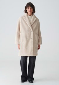 Beige, doppelt geknöpfter Teddy-Mantel mit Seitentaschen, Schalkragen und strukturiertem Stoff. Kombiniert mit schwarzen, weit geschnittenen Hosen und schwarzen Schuhen.
