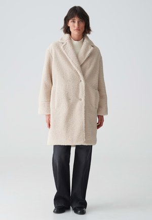 Beige teddy coat met dubbele sluiting, zijzakken, sjaalkraag en een textuurstof. Gecombineerd met zwarte wijde broek en zwarte schoenen.