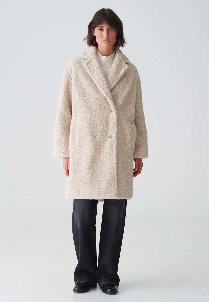 Beige, doppelt geknöpfter Teddy-Mantel mit Seitentaschen, Schalkragen und strukturiertem Stoff. Kombiniert mit schwarzen, weit geschnittenen Hosen und schwarzen Schuhen.