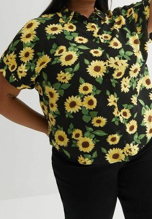 Personne portant une chemise noire boutonnée avec un motif de tournesols jaunes vifs et de feuilles vertes, associée à un pantalon noir, debout avec un bras levé.