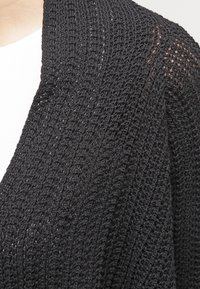 Svart stickad kofta med öppen framkant, som har en lös vävstruktur och en lätt glans. Designen inkluderar nedhasade axlar och långa ärmar.
