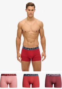 Czerwone bawełniane bokserki z szerokim czarnym pasem z napisem "SUPERDRY". Wyświetlono trzy warianty: jednolicie czerwony oraz dwa teksturowane odcienie czerwieni.