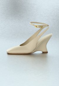 Bēša sieviešu kurpes ar kāju pacēlumu, ar smailu purngalu un potītes siksniņu ar zelta sprādzi, redzamas sānskatā uz neitrāla fona.