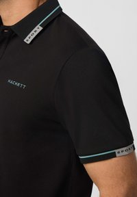 Čierna polokošeľa s krátkym rukávom a tyrkysovým textom "Hackett" na hrudi, tyrkysový pruh a štítok "SPORT" na golieri a okrajoch rukávov, nosená osobou.