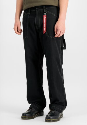 Person trägt schwarze Hose mit weitem Bein und weißen Nähten, schwarze Schuhe und einen roten "Remove Before Flight" Schlüsselanhänger, der an der Gürtelschlaufe befestigt ist.