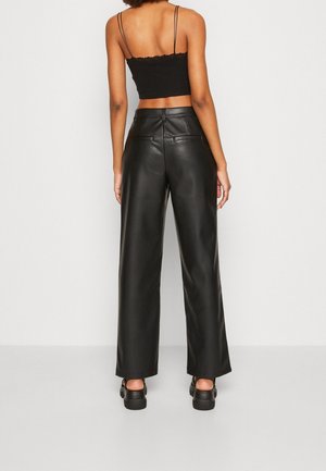 Žena stojící na bílém pozadí, oblečená v černých kožených kalhotách s širokými nohavicemi, černém krátkém topu s tenkými ramínky a černých botách na platformě.