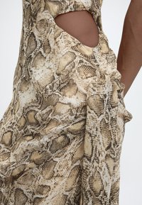 Κοντινή λήψη ενός ατόμου που φοράει φόρεμα σε μπεζ και καφέ χρώμα με φίδι σχέδιο και ένα άνοιγμα στο πλάι που αφήνει ακάλυπτο το δέρμα.