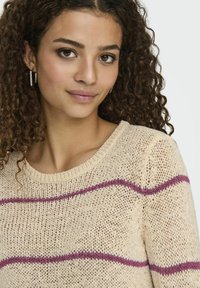 Beige gebreide trui met een gestructureerde weving, voorzien van horizontale bordeauxrode strepen. Ronde halslijn en een losse pasvorm, met subtiele ribbels bij de mouwen.