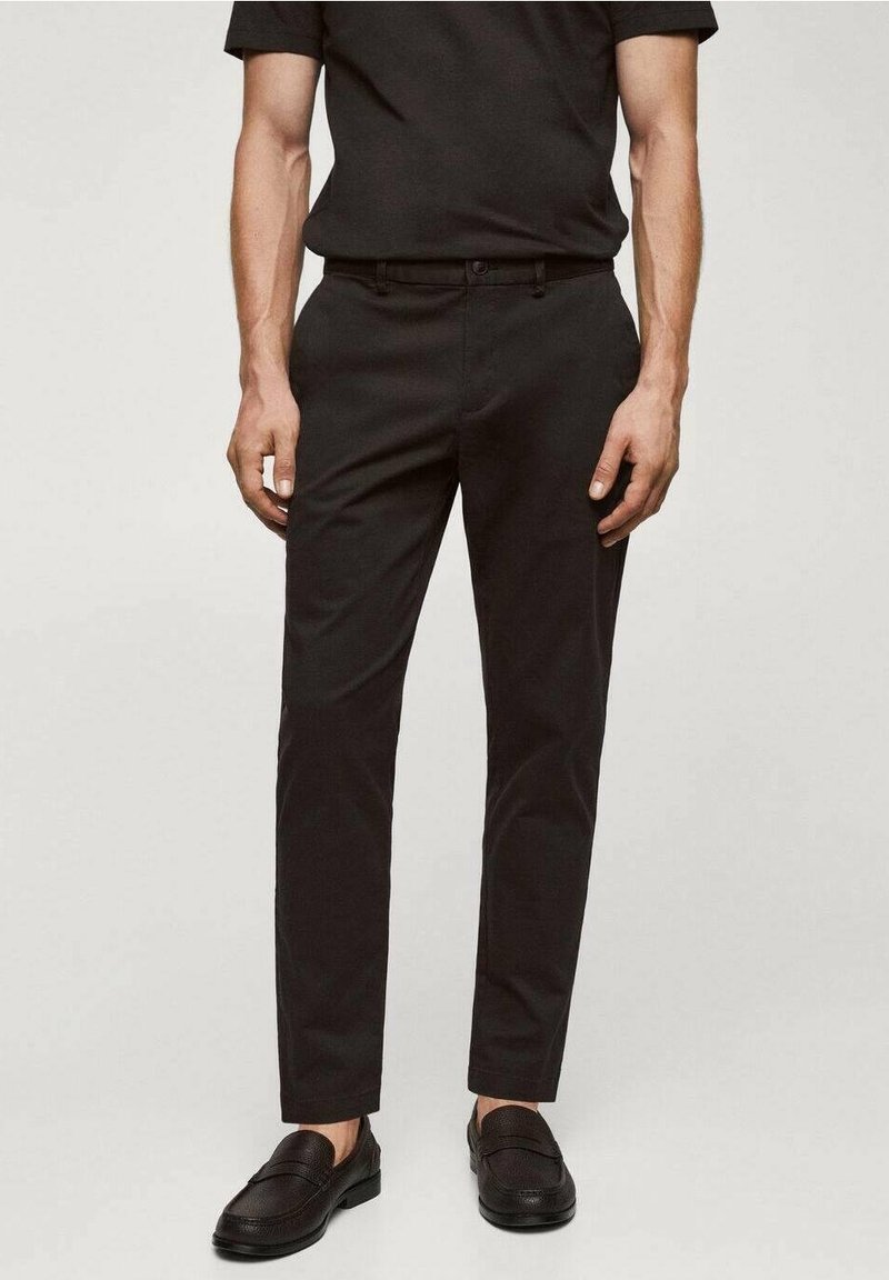 Homme portant un pantalon noir slim, des chaussures noires et une chemise noire à manches courtes, debout devant un fond clair uni.