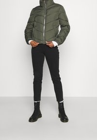 Giacca a vento verde con colletto alla coreana, chiusura frontale con zip e motivo trapuntato orizzontale. Abbinata a jeans skinny neri e stivali neri con lacci.