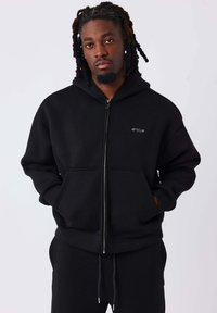 Sudadera negra con cremallera hecha de tela suave, con bolsillo canguro en la parte delantera, capucha con cordón y un pequeño logo en el pecho.