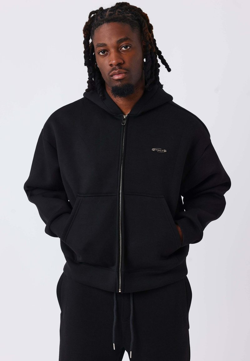 Sudadera negra con cremallera hecha de tela suave, con bolsillo canguro en la parte delantera, capucha con cordón y un pequeño logo en el pecho.