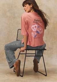 Camiseta rosa con diseño gráfico de productos de repostería, combinada con jeans azul claro y zapatos marrones, sentado en una silla de metal negra.