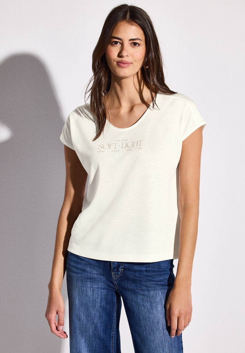 Krämfarvet kortærmet t-shirt med rund halsudskæring, prydet med en lys brun trykt tekstdesign på fronten. Sammen med blå jeans.
