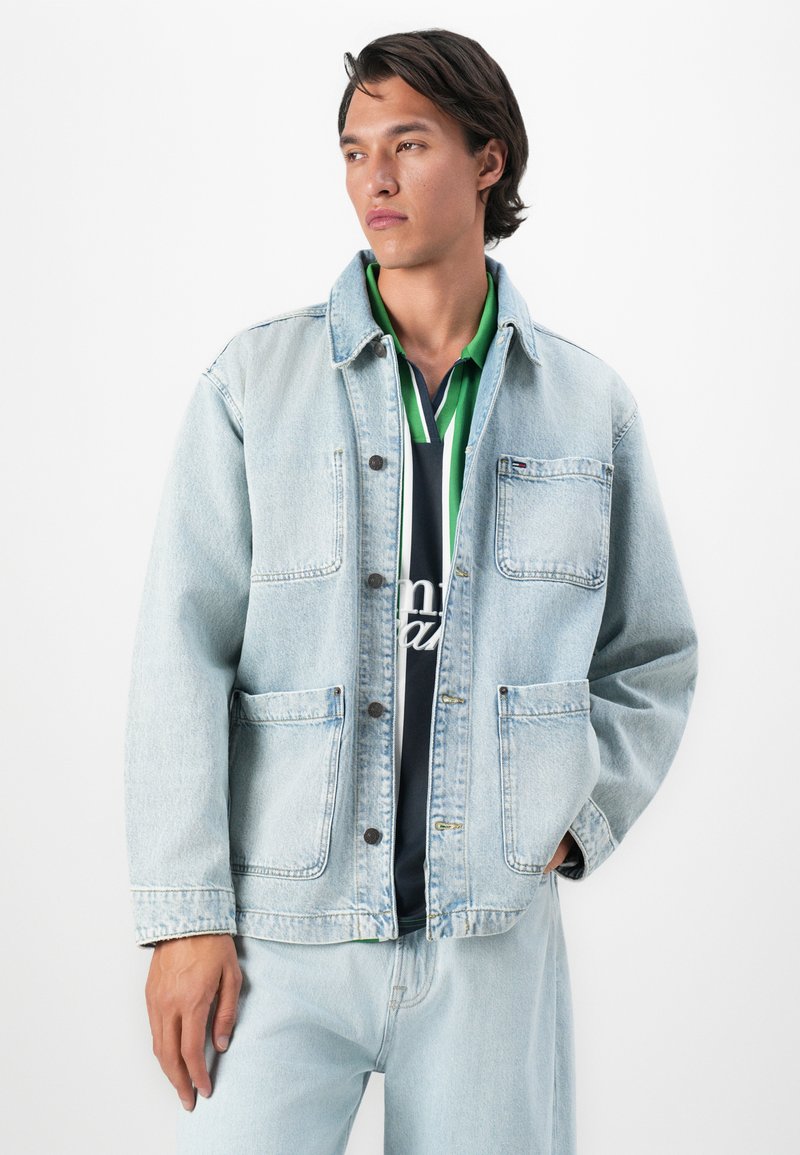 Homme aux cheveux foncés portant une veste en denim bleu clair et un jean, avec une chemise rayée verte et marine en dessous, se tenant devant un fond blanc.