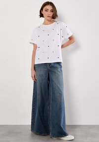Hvid kortærmet t-shirt med spredte røde, blå og sølv hjerteudskrifter. Sammen med løse blå denimjeans og hvide sneakers.