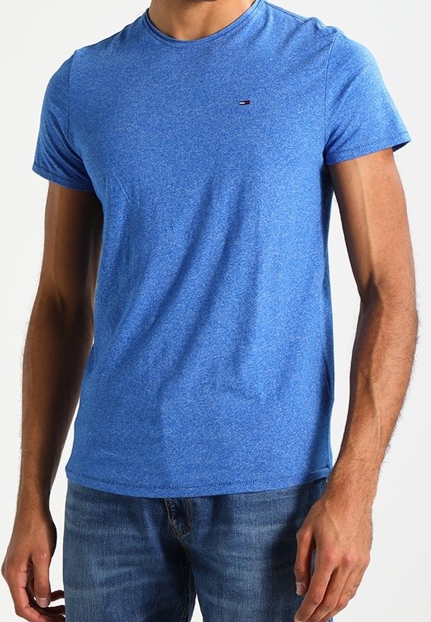 Mann in einem taillierten blauen Rundhals-T-Shirt mit kleinem Logo auf der Brust und blauen Jeans, der mit entspannten Händen an den Seiten dasteht.