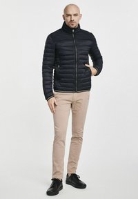 Czarna kurtka puchowa z wysokim kołnierzem, poziomym pikowaniem i kieszeniami na zamek, zestawiona z beżowymi spodniami i czarnymi trampkami.