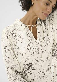 Een vrouw met krullend haar poseert, gekleed in een lichtgekleurde blouse met zwarte spetterpatronen, en laat een ontspannen en zelfverzekerde houding zien.