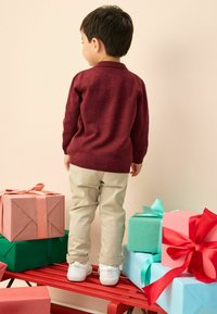 Bordeauxtrui, beige broek en witte sneakers gedragen door een kind dat op een rode houten bank staat naast kleurrijk ingepakte cadeaus.