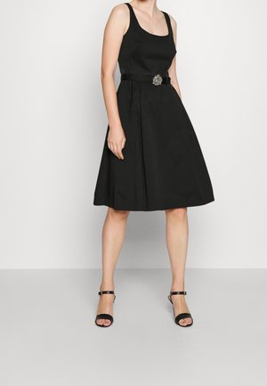 Kobieta w czarnej sukience bez rękawów z rozkloszowanym dołem i ozdobną klamrą paska, w czarnych szpilkach z paskiem wokół kostki na jednolitym tle.