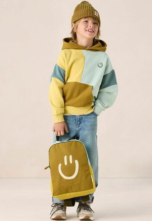 Kind dat een gele en blauwe patchwork hoodie draagt, een mosterdkleurige gebreide beanie, jeans, sneakers en een groene rugzak met een grote witte glimlach.