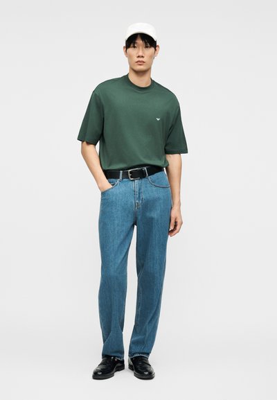 Νέος άνδρας που φοράει σκούρο πράσινο φαρδύ T-shirt, ανοιχτό μπλε ίσια τζιν, μαύρη ζώνη, μαύρα παπούτσια και άσπρο καπέλο, στέκεται με το ένα χέρι στην τσέπη.